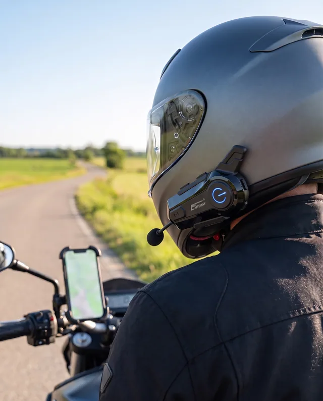 Motard vu de dos par-dessus l'épaule sur une route de campagne ensoleillée, casque intégral anthracite avec le boîtier sur le bord inférieur gauche, téléphone sur un support de guidon